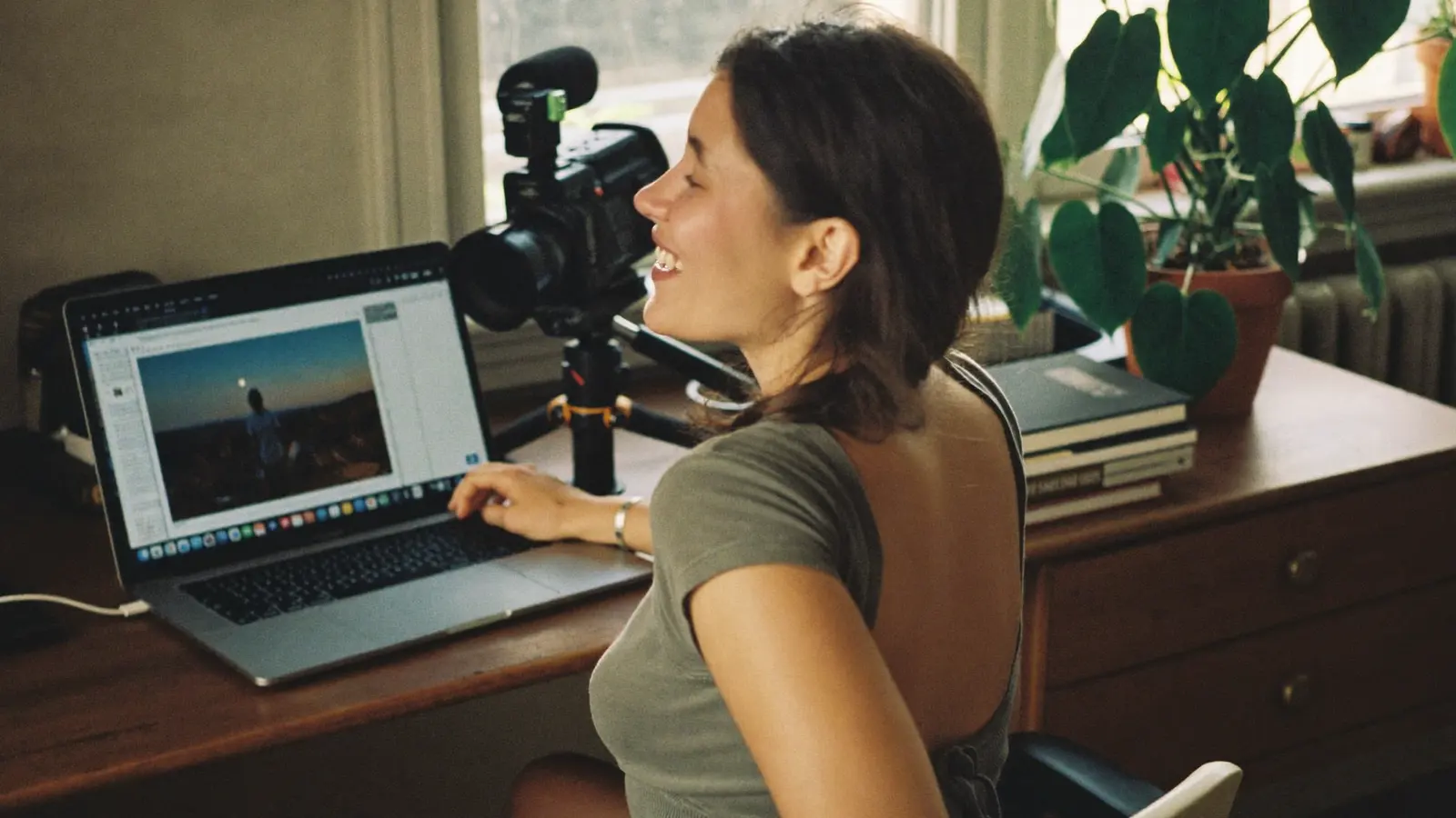 A woman at a desk in a sunlit room, laughing at something on her laptop, a camera on a tripod beside her.